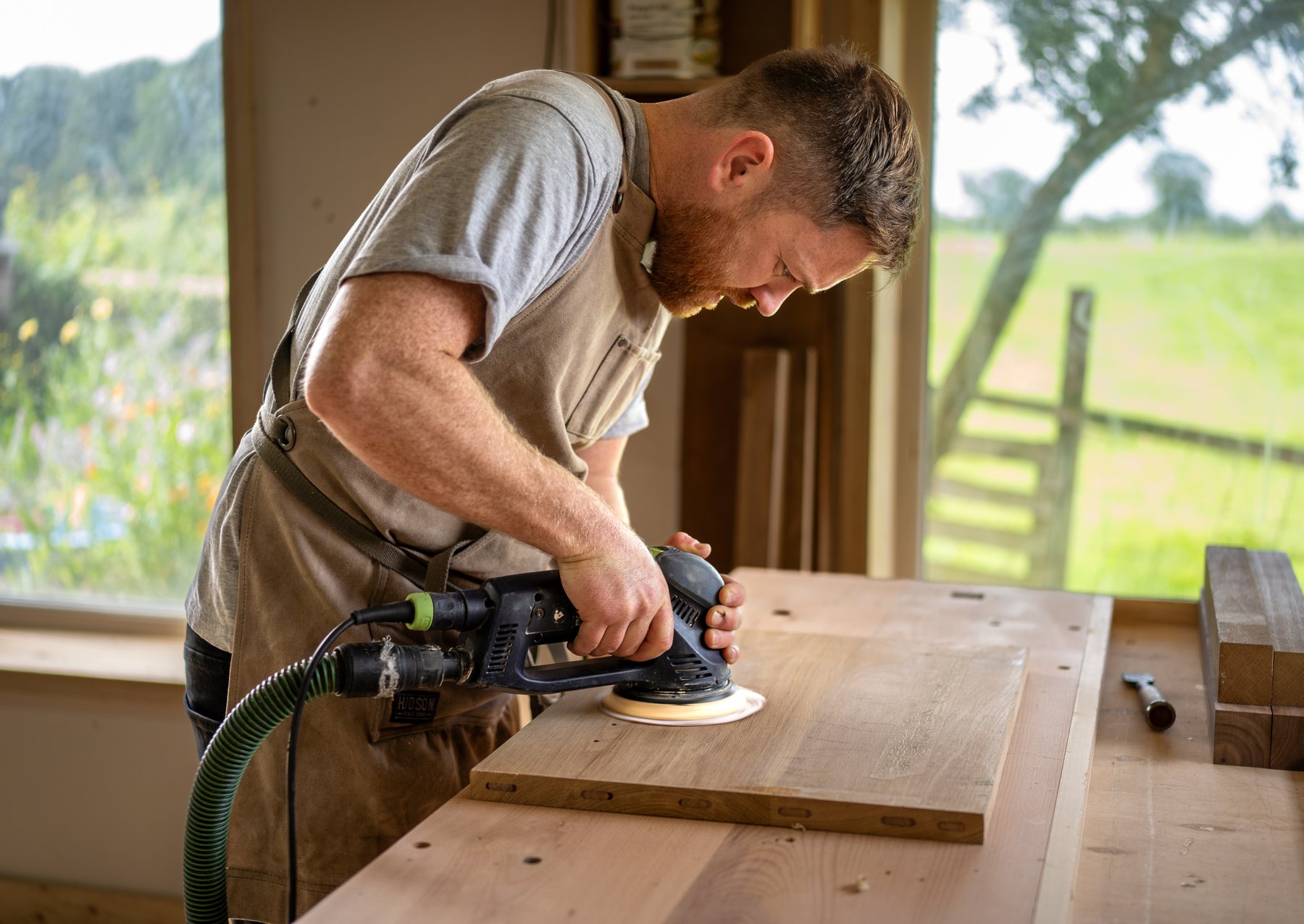 Robert Scott sanding a tabletop at the bench, fields through the window behind him.